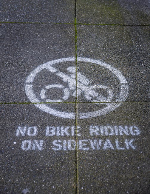 A close-up view of a paved sidewalk with textured, dark gray concrete slabs. The slabs have visible joints and some small green moss or weeds growing in the cracks. On the surface, there is a white st